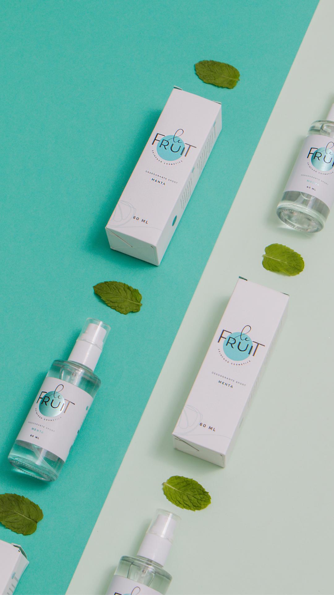 Uma fotografia exibe caixas de desodorantes Le Fruit, na variante Sport Menta, dispostas em um fundo composto por duas cores alternadas e intercaladas com folhas de menta.