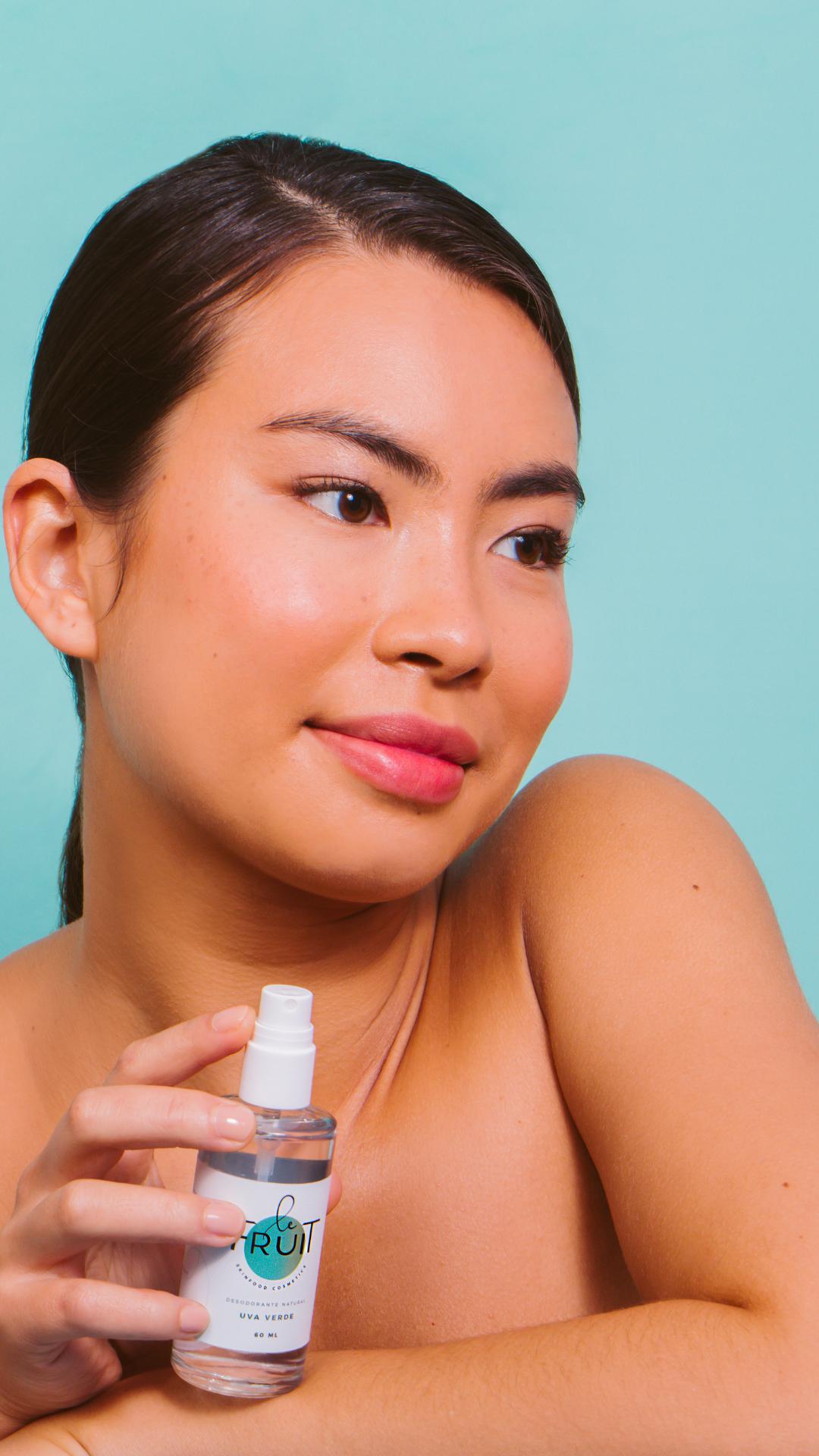 Foto em fundo azul mostra em primeiro plano o rosto de uma mulher com traços asiáticos com ombros a mostra segurando um frasco de desodorante natural