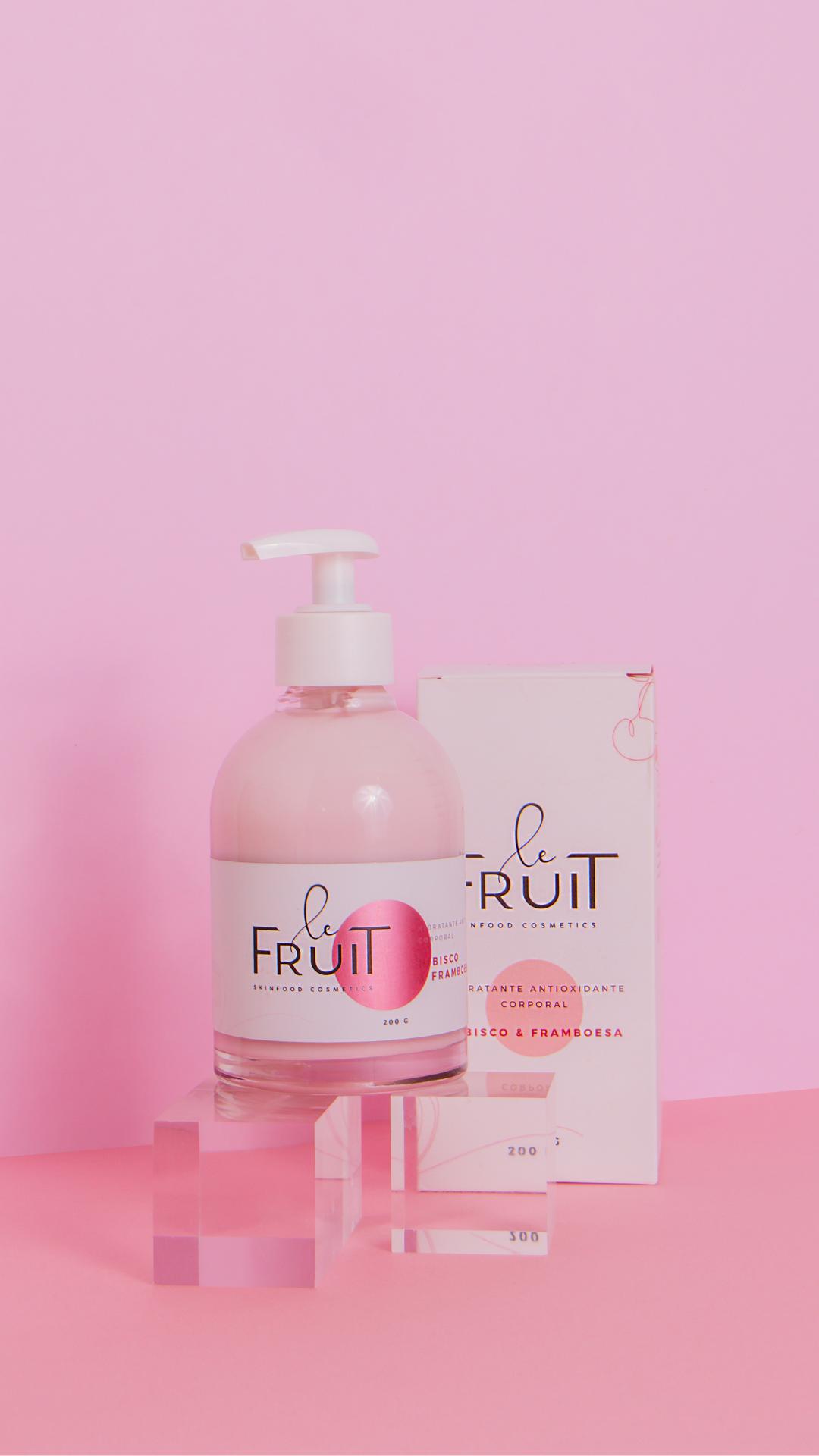 Em um fundo cor de rosa está, acima de um bloco transparente, a embalagem de hidratante da Le Fruit. Ao fundo, a caixa do produto.