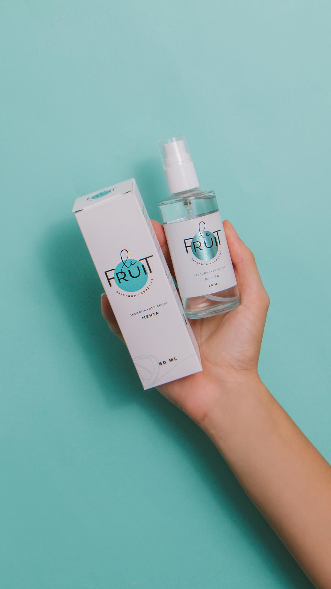 Foto em fundo azul mostrando uma mão segurando duas embalagens, uma de vidro e outra de papel, de um Desodorante Sport Menta da Le Fruit