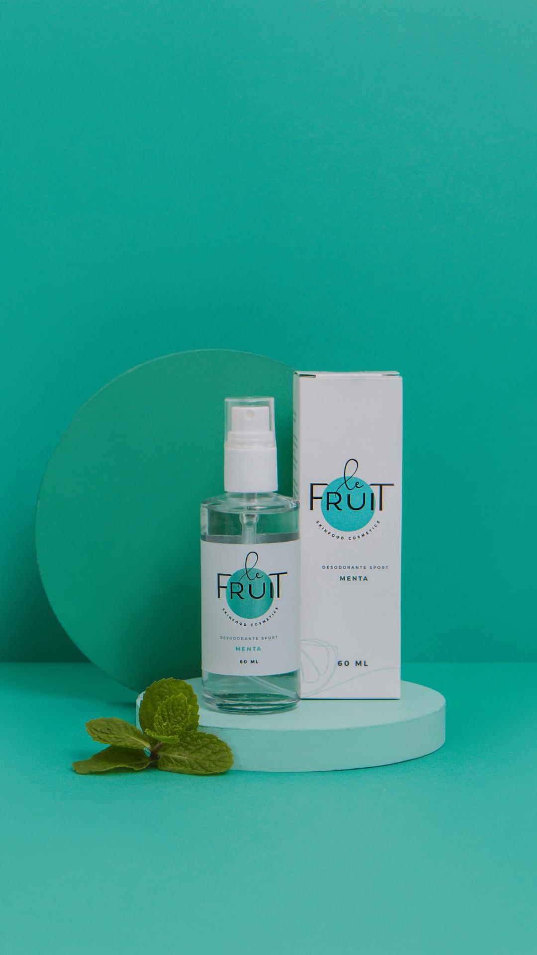 Foto das duas embalagens do Desodorante Sport Menta da Le Fruit (de vidro e de papel) uma ao lado da outra, acima de um suporte redondo e com uma folha de menta ao lado. O fundo da foto é todo azul.