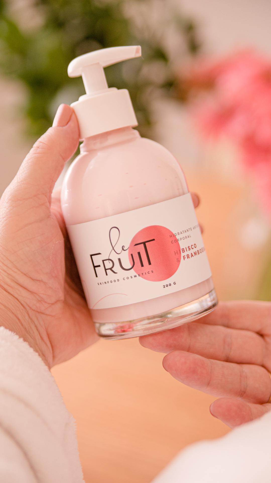 A imagem retrata duas mãos de uma mulher de pele clara segurando um frasco de vidro contendo hidratante da marca Le Fruit, cujo líquido rosa é claramente visível.