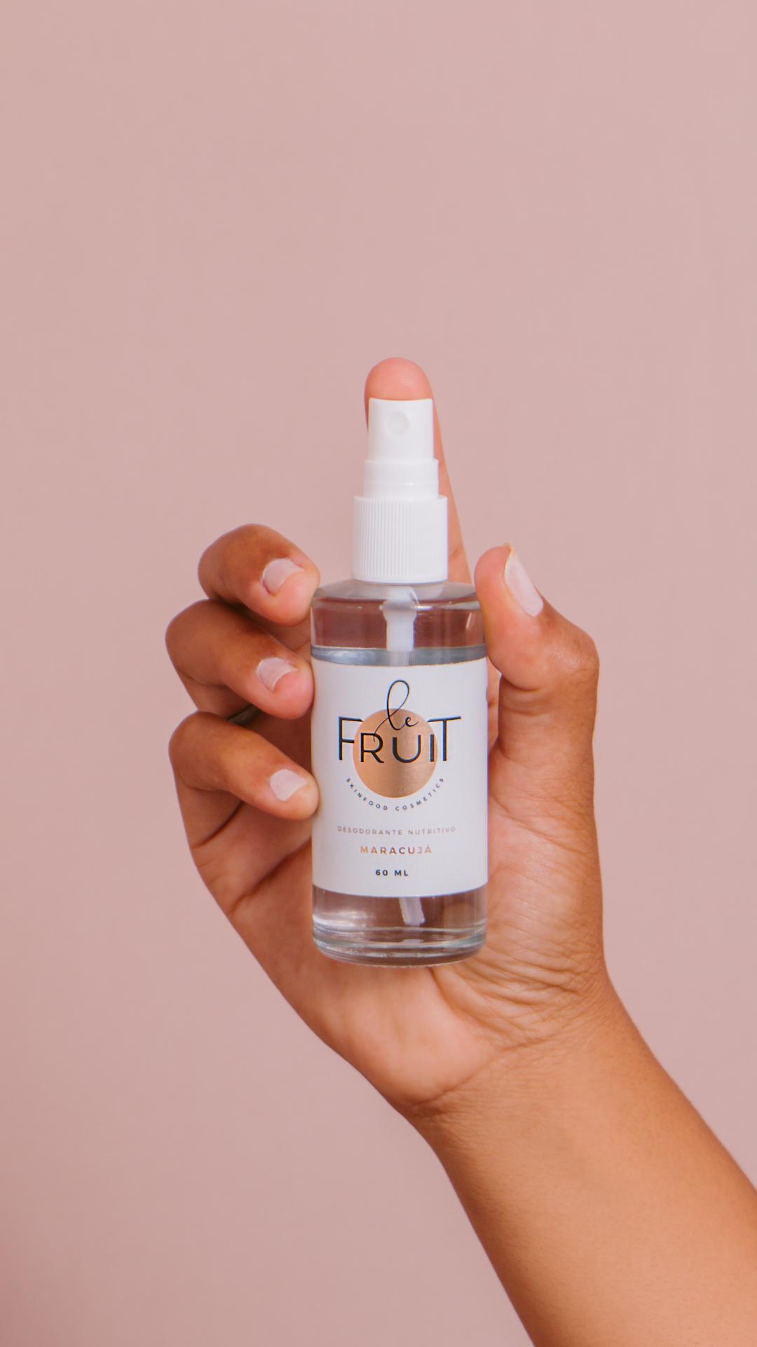 Uma embalagem de vidro do Desodorante Nutritivo Maracujá da Le Fruit é segurada por uma mão sobre um fundo rosa claro.