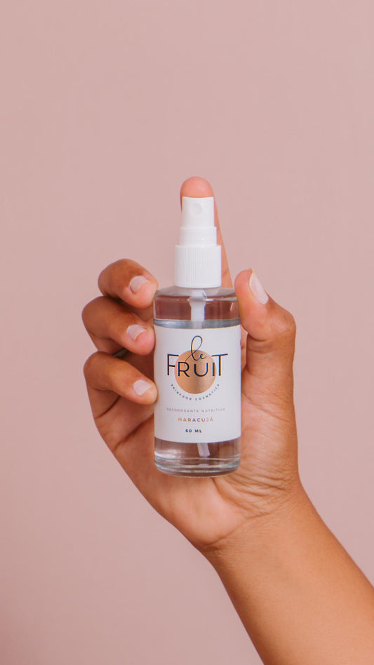Uma embalagem de vidro do Desodorante Nutritivo Maracujá da Le Fruit é segurada por uma mão sobre um fundo rosa claro.