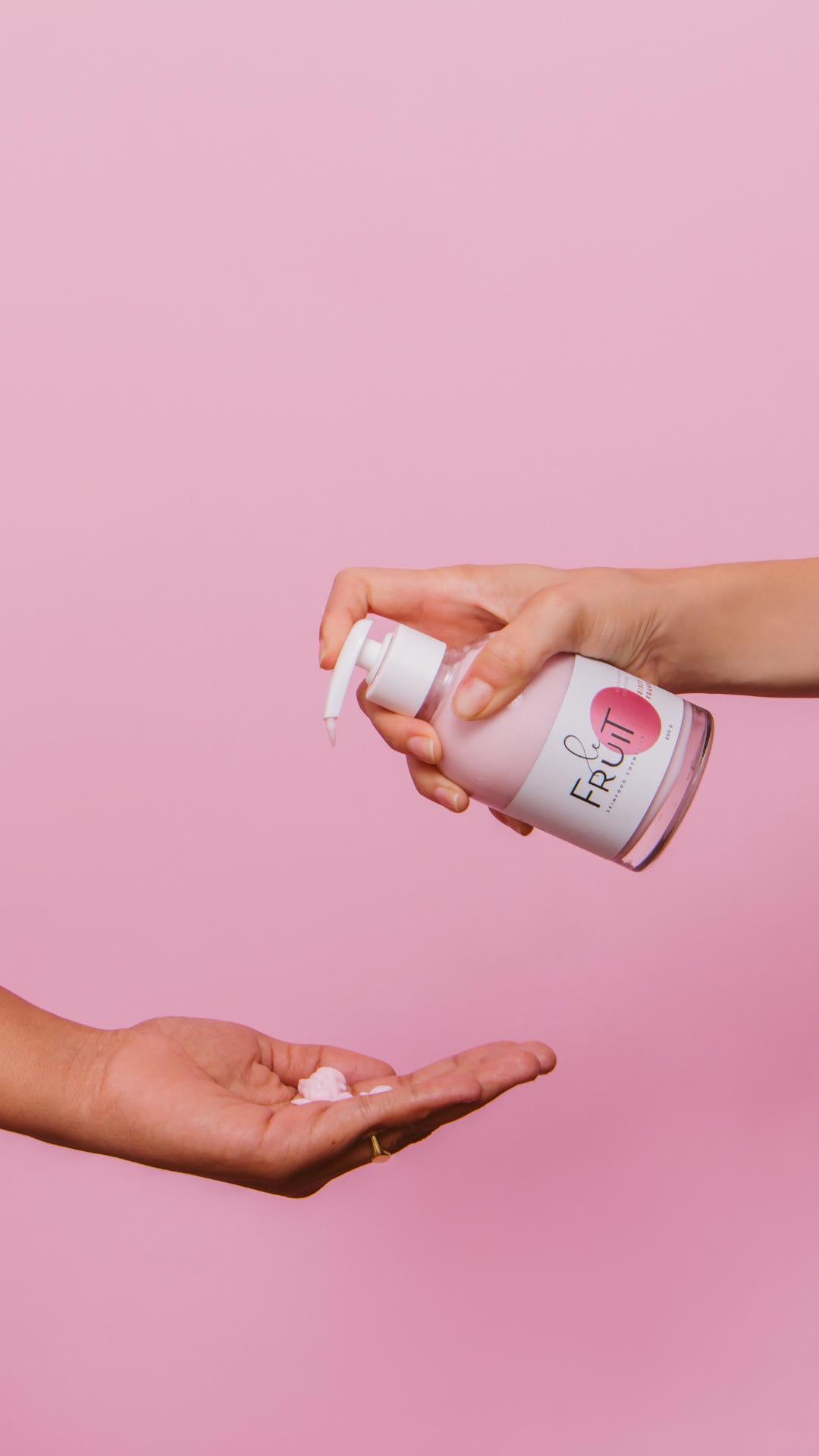 Sobre um fundo rosa, uma mão pressiona uma embalagem de vidro contendo hidratante contra outra mão, que está recebendo o creme.