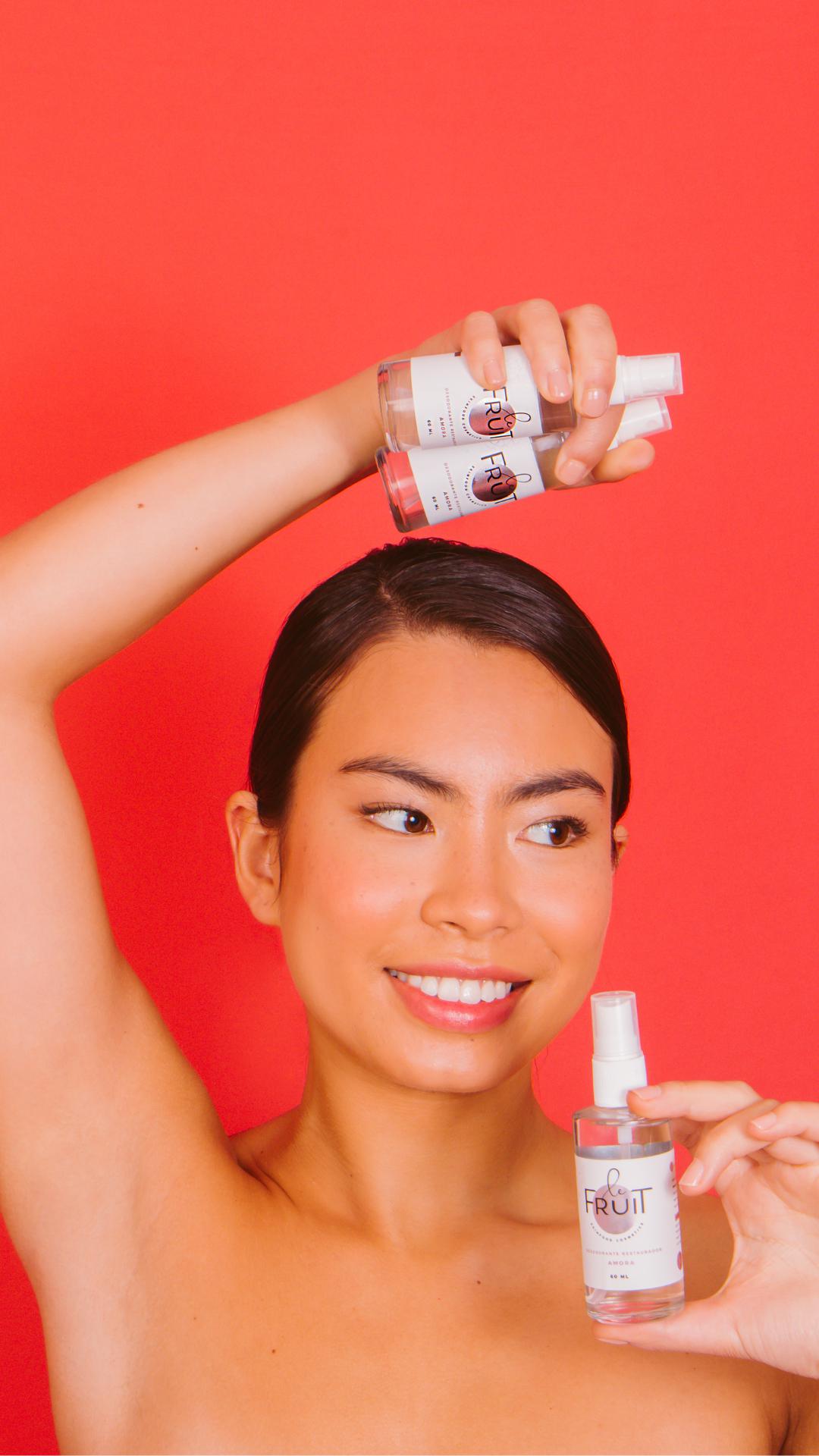 Uma mulher com características asiáticas é vista sorrindo, do busto para cima, em frente a um fundo vermelho, enquanto segura três frascos de vidro do Desodorante Le Fruit.