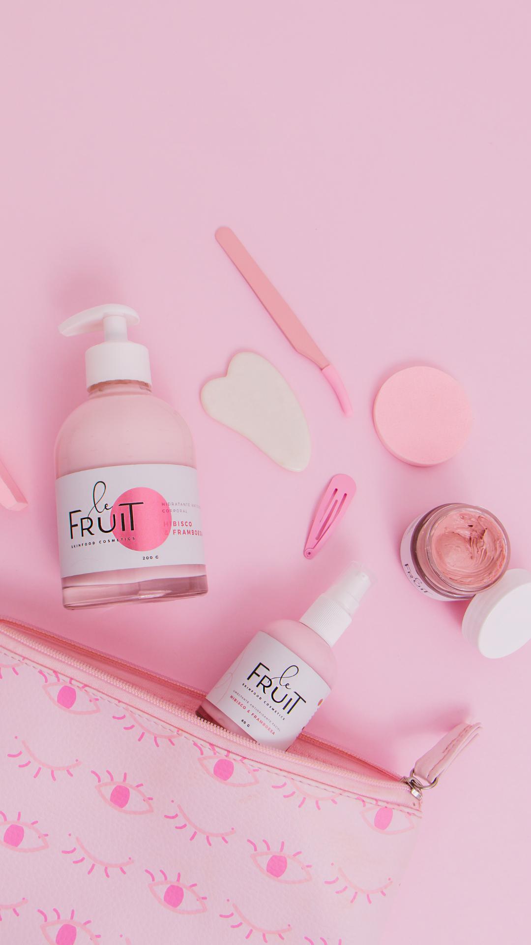 A cena retratada apresenta um fundo rosa, no qual uma necessaire rosa encontra-se aberta. Sobre ela, estão dispostos um hidratante, um umectante e uma máscara facial da marca Le Fruit, todos seguindo a tonalidade rosa.