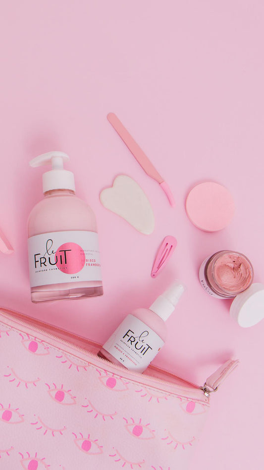 A cena retratada apresenta um fundo rosa, no qual uma necessaire rosa encontra-se aberta. Sobre ela, estão dispostos um hidratante, um umectante e uma máscara facial da marca Le Fruit, todos seguindo a tonalidade rosa.