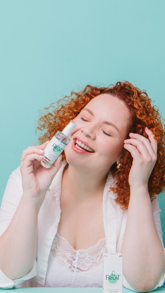 Uma mulher de pele clara e cabelos ruivos está vestindo um robe branco, sorrindo com os olhos fechados enquanto segura um desodorante de uva verde em uma das mãos.