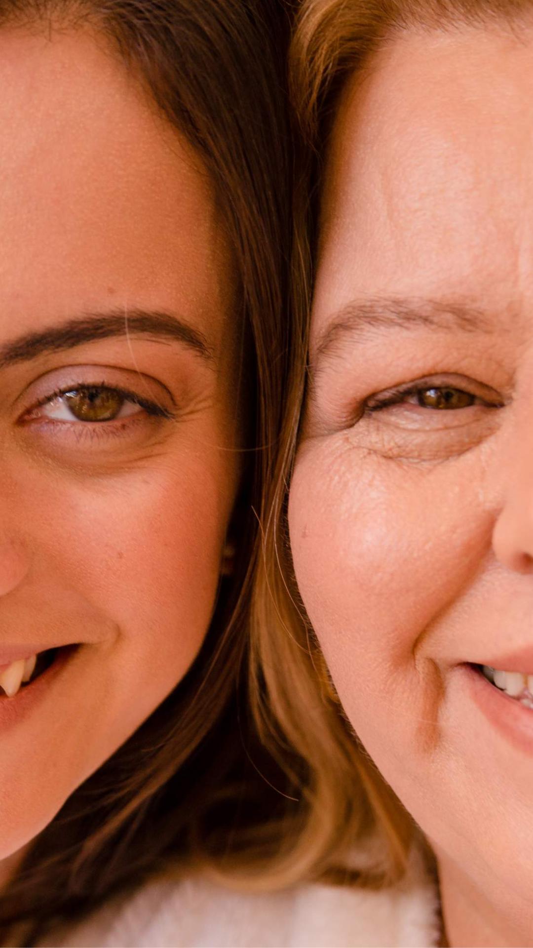 Foto de mãe e filha lado a lado, com foco no rosto sorridente de ambas e no olhar. As cabeças estão unidas e a foto mostra metade da face de casa uma.